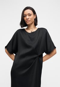 Schwarzes Kleid aus glattem Stoff, mit rundem Ausschnitt, lockeren Ärmeln und einem Knoten-Detail an einer Seite für zusätzliche Struktur.