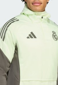 Lys grøn zip-up jakke med grå accenter, der har tre sorte striber på ærmerne og Real Madrid-logoet på brystet.