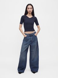 Femme debout, les mains dans les poches, portant un haut noir ajusté à manches courtes et un jean bleu à jambes larges avec une ceinture, sur un fond uni.
