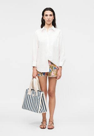Donna in piedi indossa una camicia bianca a maniche lunghe, pantaloncini floreali con bordo in pizzo, sandali marroni e tiene una grande borsa a righe blu navy e bianco.