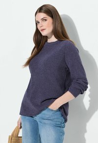 Dunkellila Pullover mit strukturiertem Strick, rundem Ausschnitt und hochgekrempelten Ärmeln, kombiniert mit hellblauen Jeans und einer gewebten Handtasche.