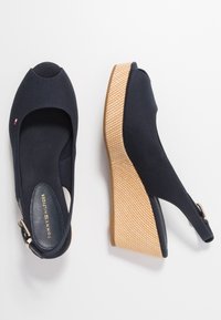 Paire de sandales compensées noires à bouts ouverts, brides réglables à la cheville et talons texturés couleur beige, marquées Tommy Hilfiger à l'intérieur.