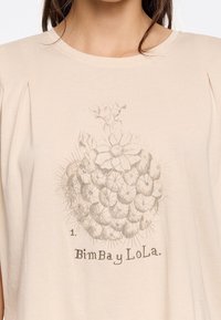 Beige short-sleeve top featuring a light gray cactus graphic with floral details. The text "1. BimBay LoLa" is printed below the design.