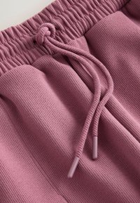 Pantalon de survêtement mauve avec texture côtelée, doté d'une taille élastique et de deux poches latérales ; les extrémités du cordon de serrage sont finies dans une couleur assortie.