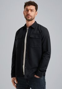 Camisa negra de manga larga con frente abotonado, dos bolsillos en el pecho y detalles texturizados en los hombros. Usada sobre una camiseta blanca.