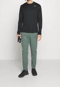 Svart långärmad träningströja, byxor med grön elastisk midja, svarta sneakers, håller i ett hopprep, med en logotyp på tröjan och byxorna.