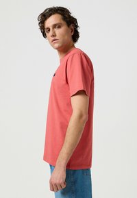 Korallfärgad bomullströja med korta ärmar, rund halsringning och en liten logodetalj på bröstet; tillsammans med blå denimjeans.