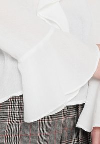 Blusa bianca dalla texture con maniche a sbuffo, abbinata a una gonna a quadri grigia con accenti rossi. Focus sui dettagli del tessuto e del design delle maniche.