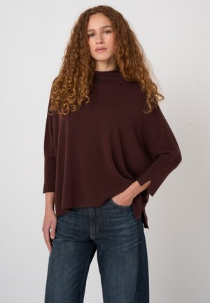 Vrouw met lang krullend haar, een losse donkerbruine trui en blauwe jeans, staand met één hand in de zak tegen een effen achtergrond.