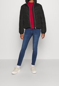 Giacca nera imbottita con cuciture a zig zag, chiusura con zip frontale e colletto alto, abbinata a un maglione rosso e jeans skinny blu. Sneakers bianche.