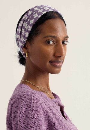 Vrouw met donker haar en een bloemenpaars hoofdband, die gouden oorringen, gouden kettingen en een paars gebreide trui met textuur draagt, zijwaarts gekeerd.