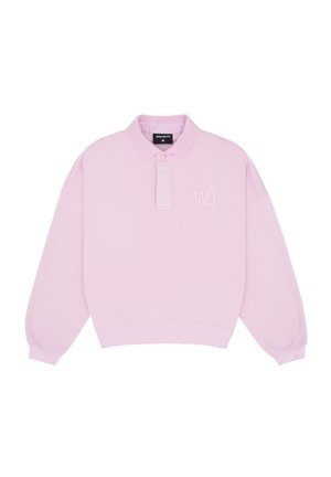 Femme portant un polo rose oversize, un short gris, des sandales marron à talons, tenant une casquette rose assortie et portant des lunettes, debout devant un fond uni.