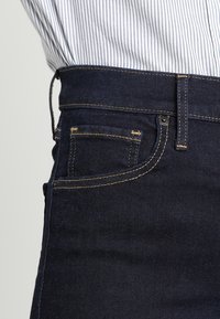 Primer plano del bolsillo y la pretina de unos vaqueros de mezclilla azul oscuro con costuras amarillas, llevados con una camisa blanca metida con finas rayas azules.