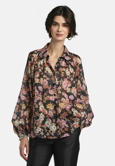Donna che indossa una blusa nera trasparente a maniche lunghe con stampa floreale rosa, gialla e bianca, abbinata a pantaloni neri e orecchini a cerchio.