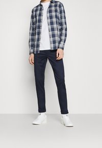 Camicia a maniche lunghe a quadri blu navy sopra una maglietta bianca, abbinata a pantaloni slim blu navy e sneaker bianche. La camicia presenta dettagli con bottoni e colletto.