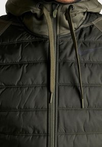 Uomo che indossa un giubbotto imbottito con cappuccio di colore verde oliva scuro, dotato di zip e lacci, con il logo Nike nero visibile sul petto.