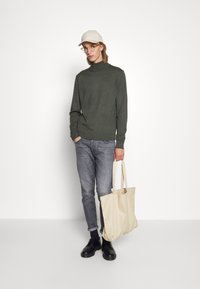 Grønn høyhalset genser, grå jeans, svarte sko og beige totebag. Tilbehør inkluderer en beige caps og briller med klare rammer.