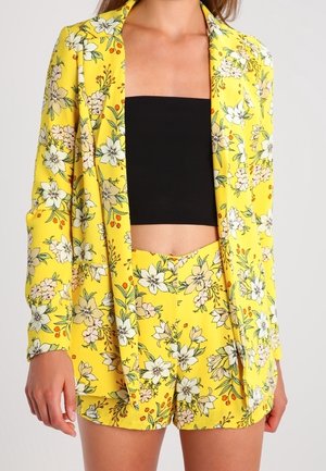 Blazer floral amarillo y shorts a juego de cintura alta combinados con un top tipo tubo negro, usados por una persona que está de pie contra un fondo liso.