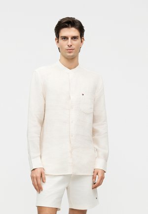 Młody mężczyzna noszący kremową, zapinaną na guziki koszulę z długim rękawem i kieszonką na piersi oraz pasujące krótkie spodenki, stojący na jednolitym tle.