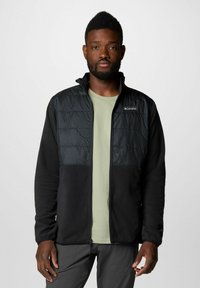 Giacca in pile nera con una parte superiore in nylon lucido trapuntato di colore nero. Presenta un collo alto e una zip, abbinata a una camicia verde chiaro sotto.