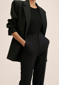 Personne portant un blazer noir avec manches retroussées, un haut noir ajusté et un pantalon noir taille haute, les mains dans les poches.