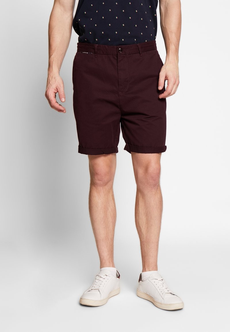 Homme portant un short bordeaux foncé à ourlet roulé, une chemise noire à motifs et des baskets blanches, debout devant un fond clair uni.