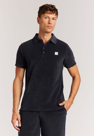 Marineblaues Poloshirt aus weichem Stoff, mit Kragen, drei Knöpfen in der Knopfleiste und einem kleinen Logo-Patch auf der linken Brustseite. Kurze Ärmel.