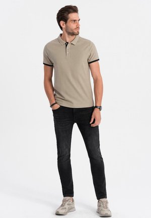 Beige polo shirt met zwarte afwerking, voorzien van een kraag en drie knopen, gecombineerd met zwarte jeans en lichte sneakers.