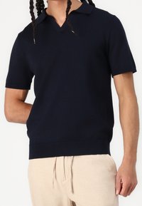 Marinblå stickad polo med V-ringning och korta ärmar, med ribbat nederkant och slät yta. Bärs med ljusbeige byxor.