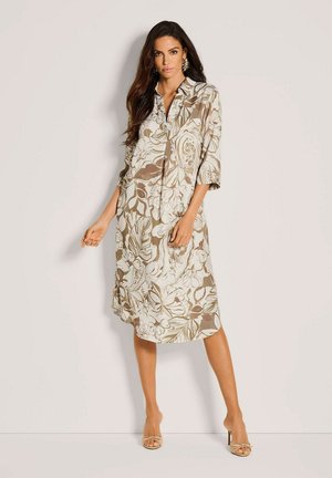 Frau trägt ein knieumspielendes Kleid in Beige und Weiß mit Blumenmuster, 3/4-Ärmeln und beige offenen Absatzschuhen, steht vor einem schlichten hellen Hintergrund.