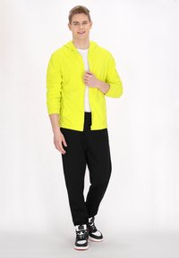 Felpa gialla neon con zip, maniche lunghe, texture morbida, abbinata a pantaloni neri e sneaker bianco e nero. Design semplice senza motivi.