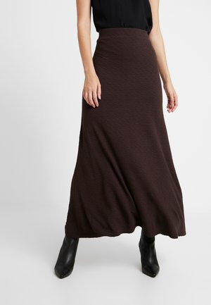 Kobieta ubrana w teksturowaną ciemnobrązową spódnicę sięgającą kostek, czarny top bez rękawów oraz czarne botki z zaostrzonym noskiem, stojąca na białym tle.