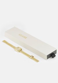 Montre en métal doré avec un cadran rectangulaire et un bracelet en maille, présentée à côté d'une boîte blanche avec "CLUSE" embossé en doré.