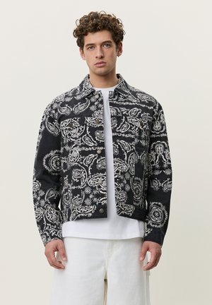 Ung mand med krøllet hår iført en sort og hvid jakke med paisleymønster over en hvid T-shirt og hvide bukser, stående foran en ensfarvet baggrund.