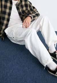 Chemise en flanelle à carreaux noirs et beiges, pantalon cargo blanc, baskets noires, assis sur un tapis bleu, main reposant sur le genou.