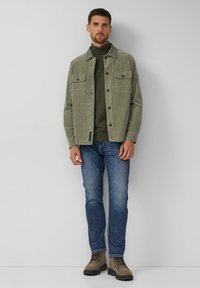 Kurtka sztruksowa w oliwkowym kolorze z dwoma kieszeniami na piersi, zestawiona z ciemnozielonym golfem, niebieskimi jeansami i beżowymi sznurowanymi botkami.
