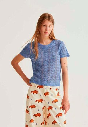 Mujer joven con un suéter de punto azul de manga corta y pantalones crema con patrones de conejos naranja y corazones azules, de pie contra un fondo blanco.