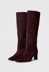Bottes hauteur genou bordeaux en matériau doux et texturé avec un bout pointu et un talon carré. Présente un design lisse et sans couture.
