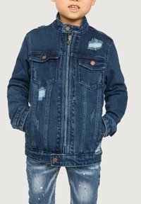 Veste en denim bleu foncé avec un col haut, des détails usés, deux poches poitrine et des accents métalliques. Portée sur un jean délavé.