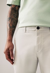 Pantaloni grigio chiaro con una texture liscia, caratterizzati da un singolo bottone marrone in vita. Accompagnati da un top verde chiaro con una texture.