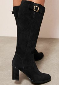 Juodos nubuko aukštakulnės batų iki kelių su blokuoto kulno ir auksiniu sagties akcentu šone. Lygus tekstūra ir pritaikytas dizainas.