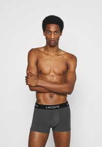 Shorts de boxe gris en tissu doux avec une ceinture élastique noire arborant le logo "LACOSTE" en blanc. Le modèle est torse nu avec les bras croisés.