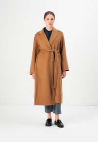 Donna in piedi di fronte, indossa un lungo cappotto color cammello con la cintura annodata, jeans grigi tagliati, mocassini neri e una camicia scura con colletto.