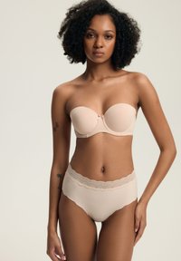 Kobieta z kręconymi włosami ubrana w stanik bez ramiączek w odcieniu nude oraz dopasowane majtki z koronkowym wykończeniem, stojąca na tle jasnego jednolitego tła.