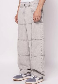 Vyriškis, vilkintis plačiais, šviesiai pilkais džinsais su įvairiaspalviais įsiuvais ir pilkai-baltomis sportbačiais, stovi prieš paprastą fono.