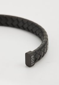 Bracciale in pelle nera intrecciata con un'estremità quadrata su cui è inciso "BOSS". La texture è liscia e piatta, con una forma curva continua.