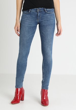 Kvinde iført tætsiddende blå jeans og skinnende røde ankelstøvler med hæl, står mod en ensartet lys baggrund.