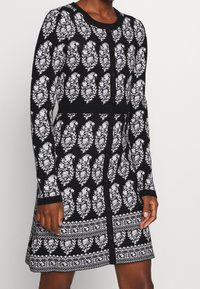 Robe en tricot noir et blanc avec motif de feuilles, manches longues, encolure ronde, fermeture par boutons et détails texturés à la taille et à l'ourlet.