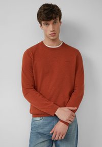 Rostfarbener Strickpullover mit klassischem Rundhals, gerippten Bündchen und Saum, getragen über einem weißen Rundhals-Shirt. Einfaches Logo auf der Brust.