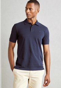 Marineblå bomullspolo med klassisk krage og korte ermer. Har en subtil logo på brystet. Matchet med lyse beige bukser.